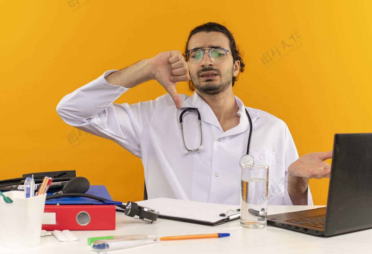 下困惑的年轻男医生戴着医用眼镜 穿着医用长袍 听诊器坐在办公桌旁笔记本电脑拇指坐着