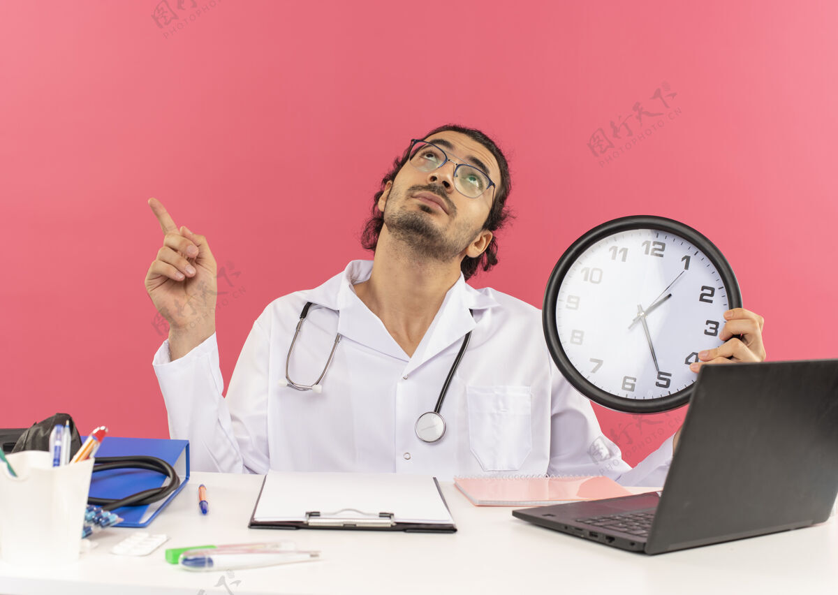 工作年轻的男医生戴着医用眼镜 穿着医用长袍 带着听诊器 坐在办公桌旁看着我男性向上工具