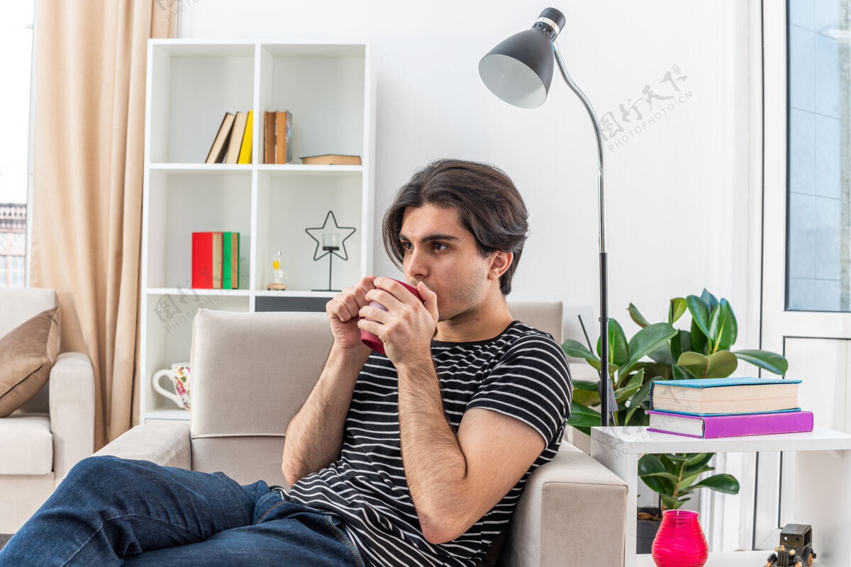 茶在明亮的客厅里 一个穿着休闲服的年轻人坐在椅子上喝着杯子里的热茶杯子热房间