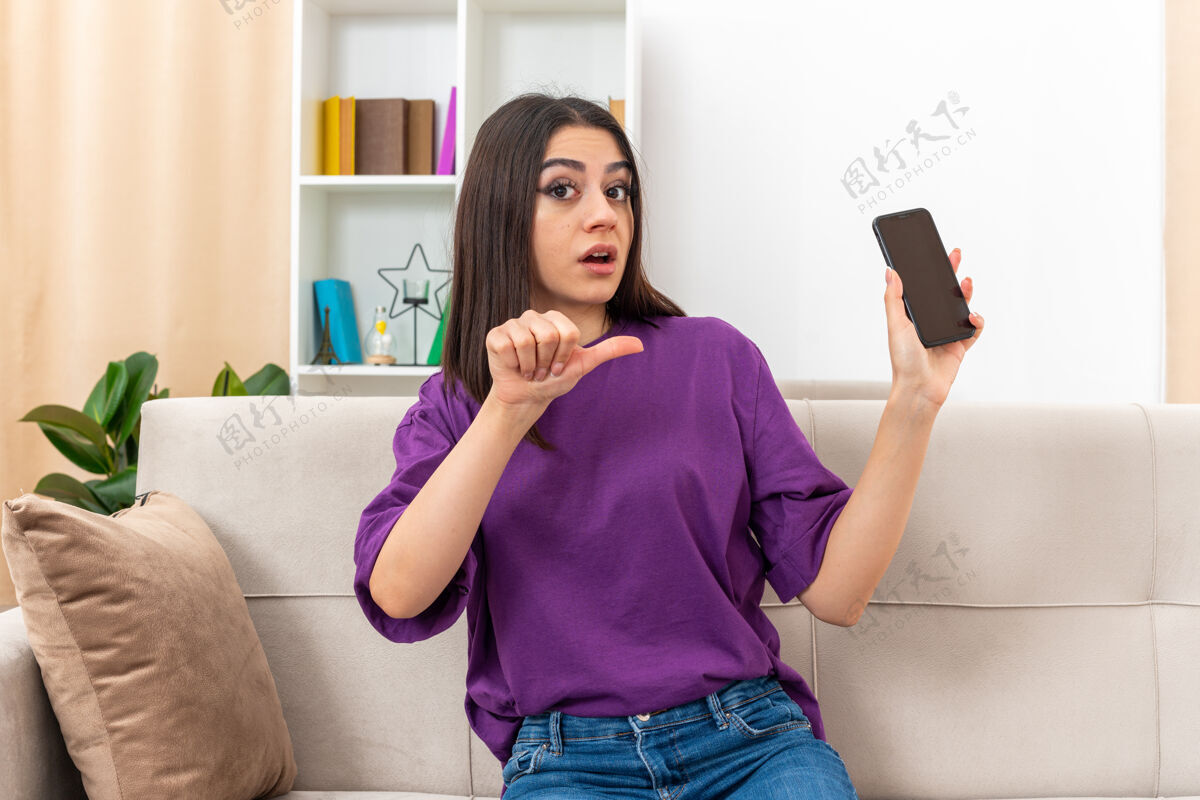 手机穿着休闲服的年轻女孩拿着智能手机 看起来很困惑 用拇指指着坐在沙发上的手机 客厅里光线明亮拇指随意坐着