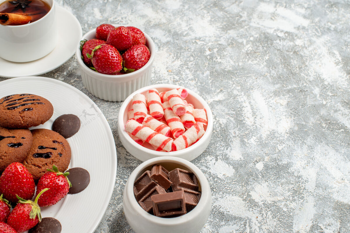 盘子下半部分在灰白色桌子的左侧 看到椭圆形盘子上的饼干草莓和圆形巧克力碗里的糖果草莓巧克力和肉桂茶肉桂巧克力椭圆形