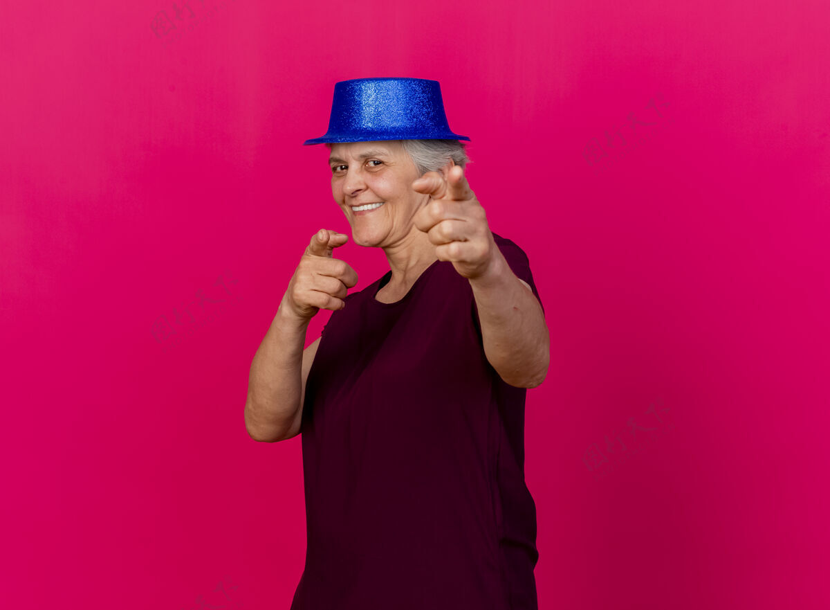 女人微笑着戴着派对帽的老妇人看着镜头 两手放在粉红色的衣服上手老人帽子