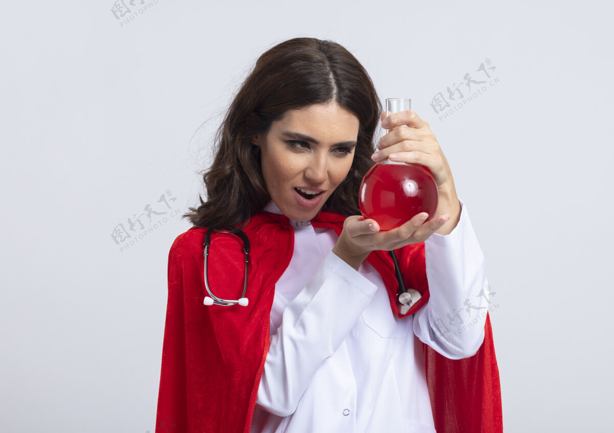 听诊器兴奋的白人超级英雄女孩穿着医生制服 戴着红色斗篷和听诊器化学斗篷液体