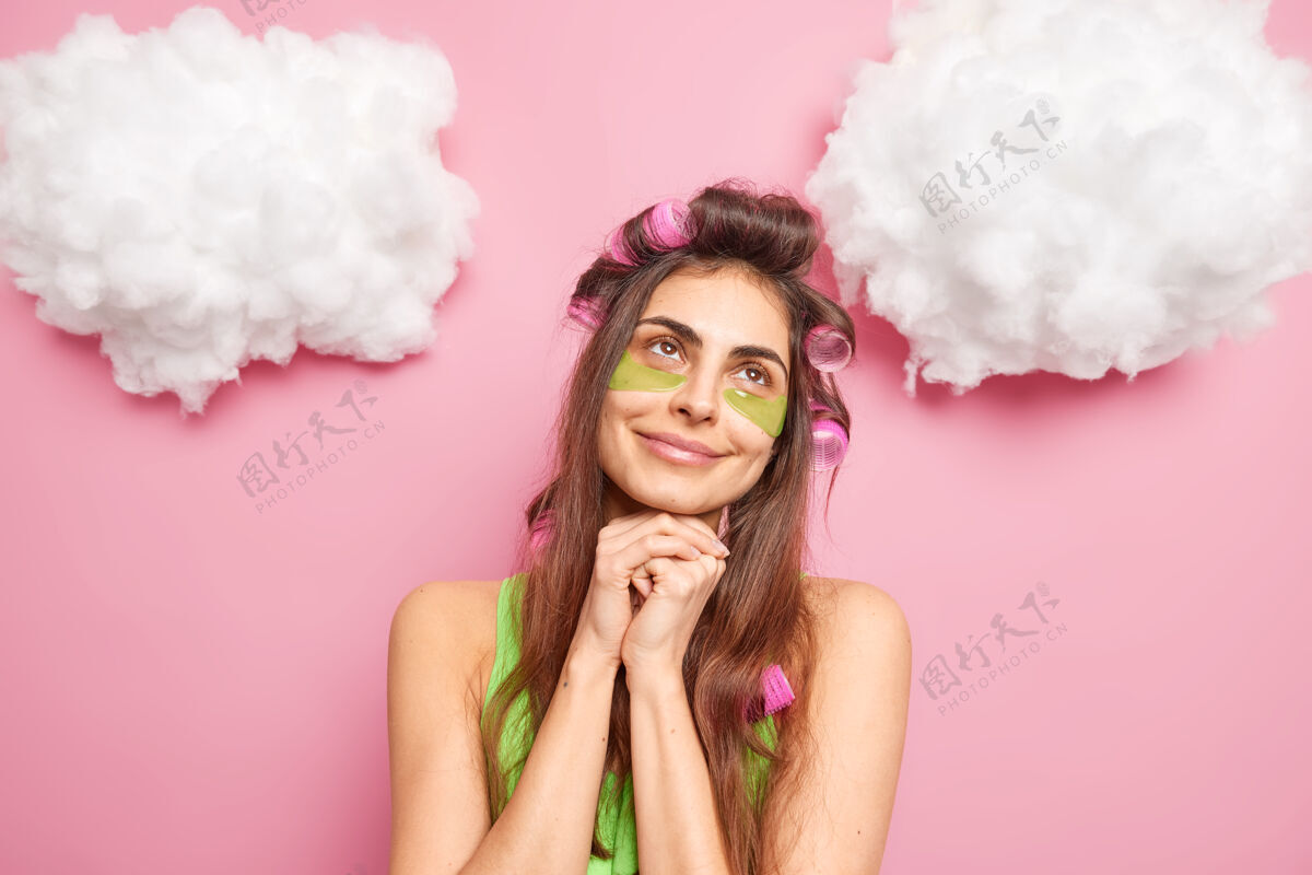 快乐喜出望外的黑发女人手放在下巴下面涂绿色胶原蛋白贴贴头发卷让发型孤立在粉色的墙壁上白云上面美容护理思考