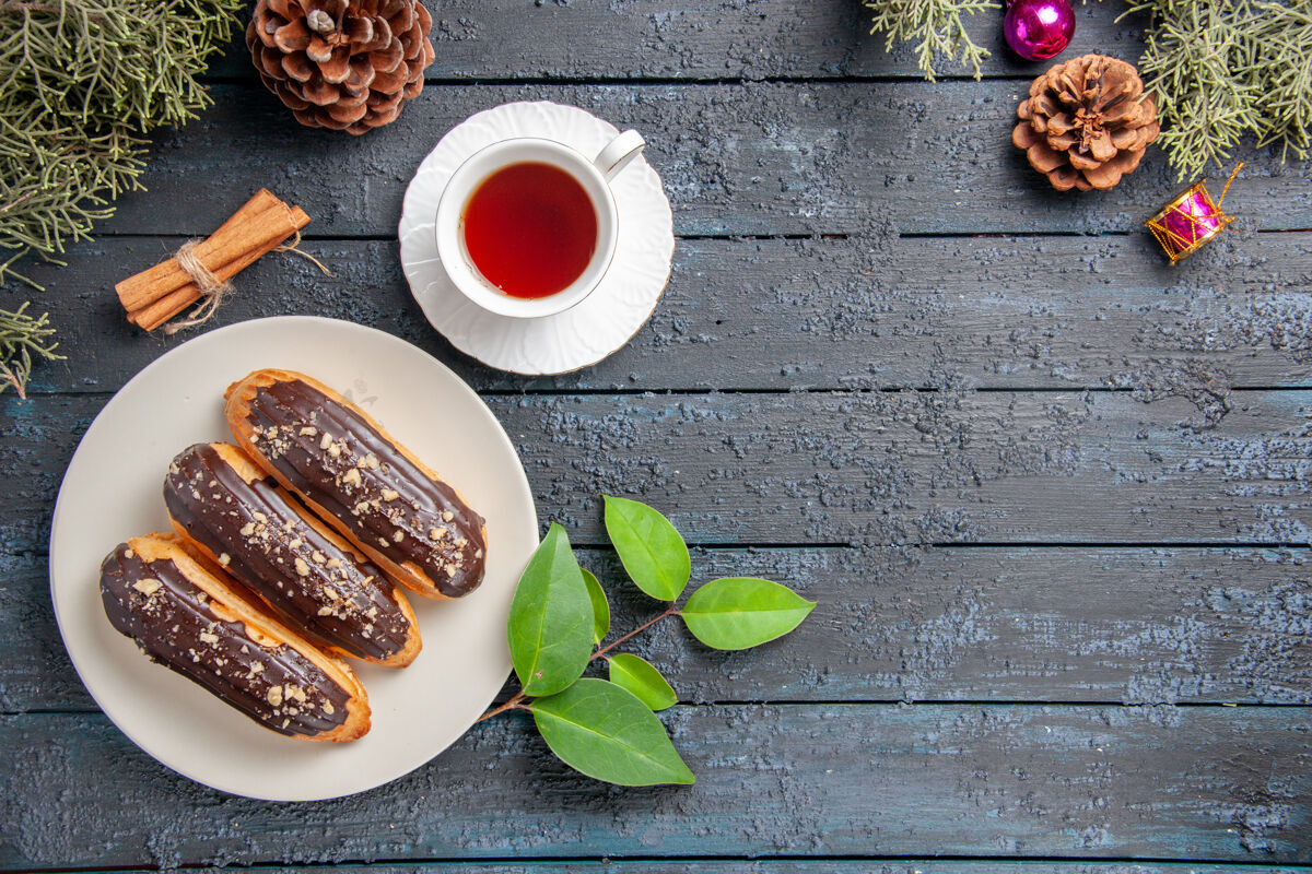 肉桂俯视图白色椭圆形圆锥体上的巧克力蛋糕圣诞玩具枞树树叶肉桂和一杯茶在黑色木质地面上复制空间午餐顶部枞树