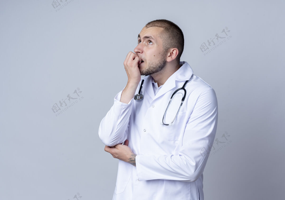 手指体贴的年轻男医生 穿着医用长袍 脖子上戴着听诊器 咬着手指 孤零零地仰望着白墙上表情脸人