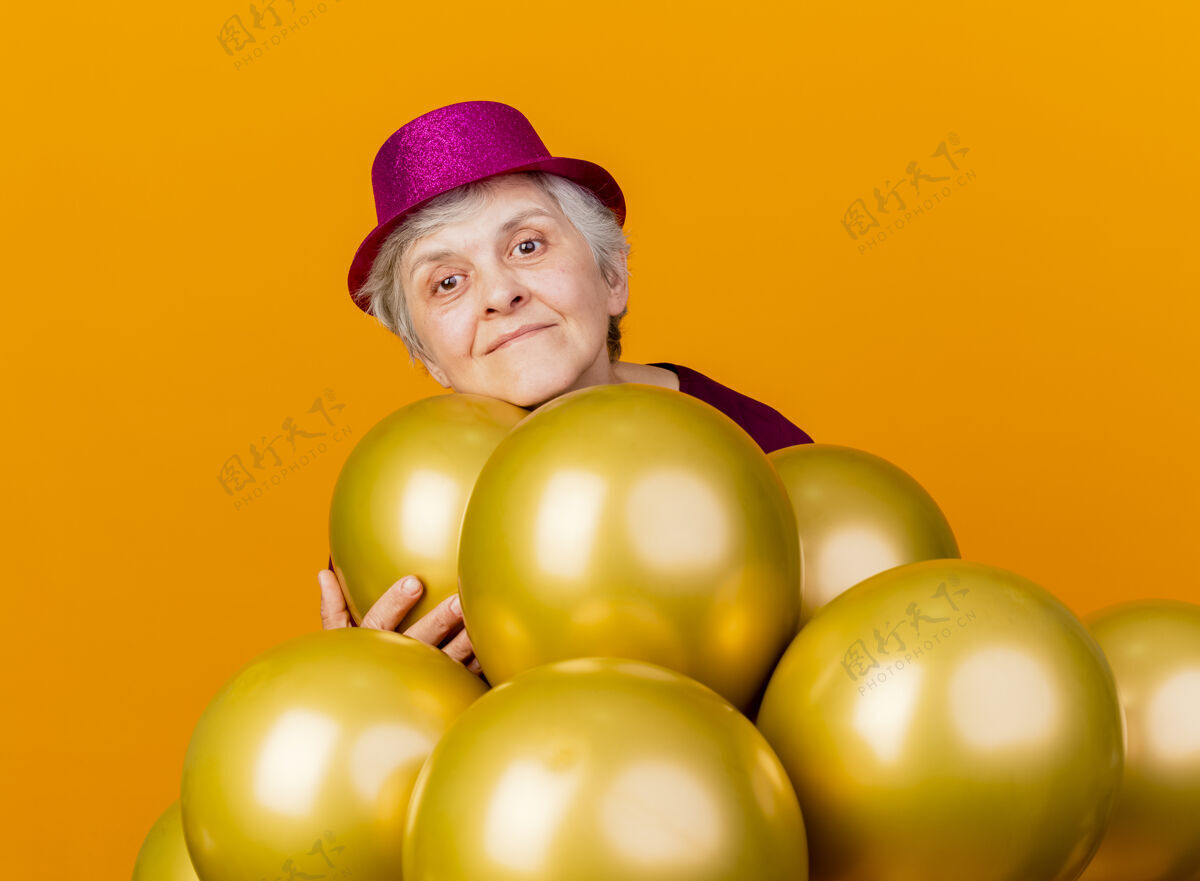 老人高兴的老妇人戴着聚会帽 手持氦气球站在橘子上穿上女人帽子