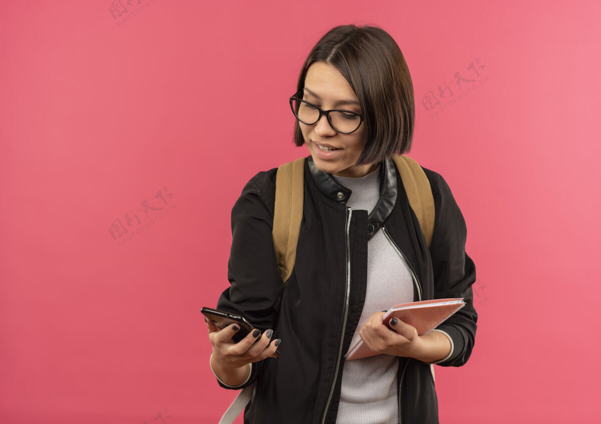 微笑微笑的年轻学生女孩戴着眼镜 背着书包 拿着笔记本和手机 看着隔离在粉红色墙上的手机学生脸垫