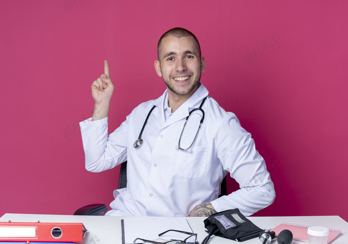 感觉面带微笑的年轻男医生穿着医用长袍 听诊器坐在办公桌旁 工作工具孤立地指向粉红色的墙上脸长袍听诊器