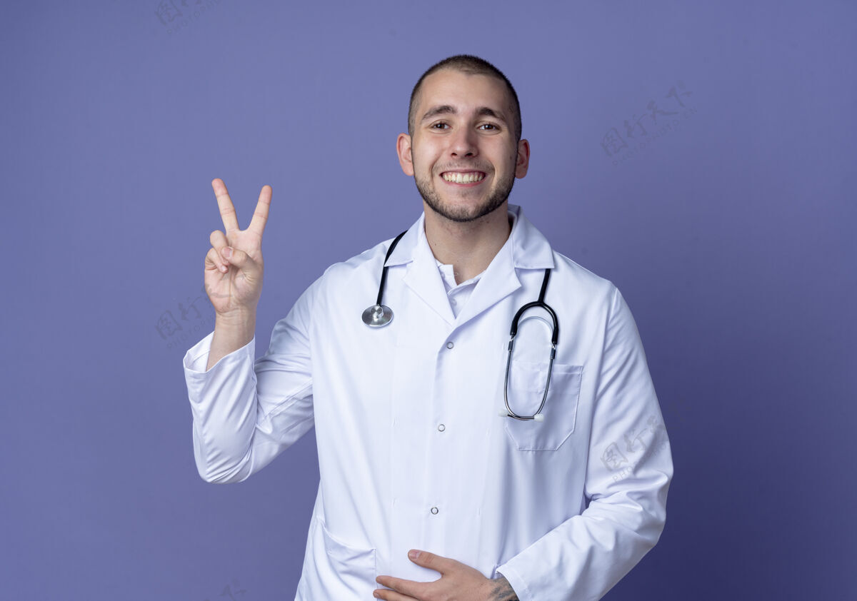 和平面带微笑的年轻男医生穿着医用长袍 戴着听诊器 做着和平的手势 手放在隔离在紫色墙上的肚子上长袍人腹部