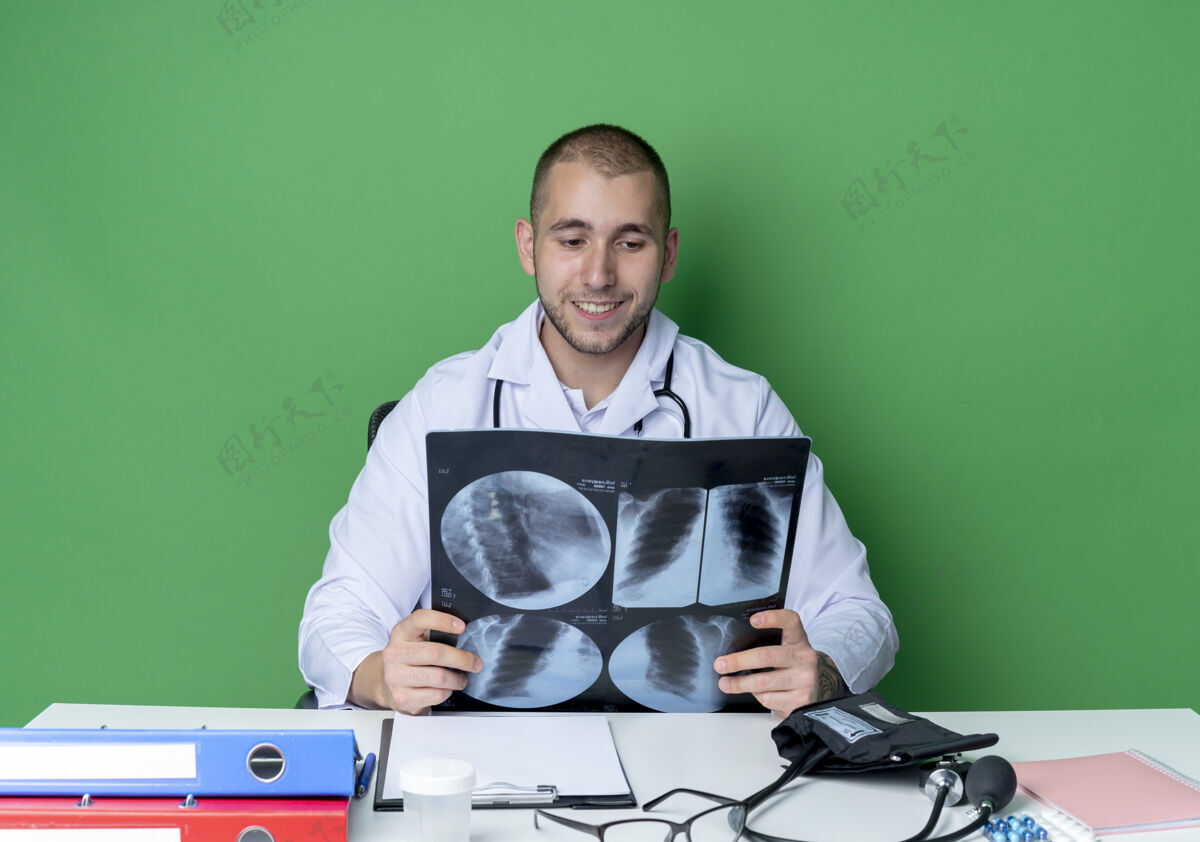 姿势面带微笑的年轻男医生穿着医用长袍 听诊器坐在办公桌旁 手里拿着工作工具 看着隔离在绿墙上的x光片抱着公民长袍