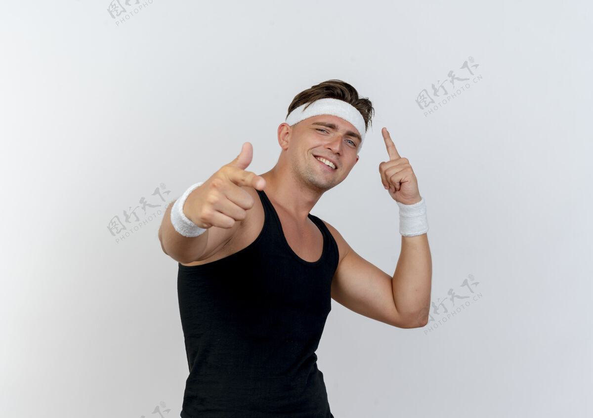 人面带微笑的年轻帅气的运动型男人戴着头带和腕带指着前面 孤立在白色的墙上白头带腕带