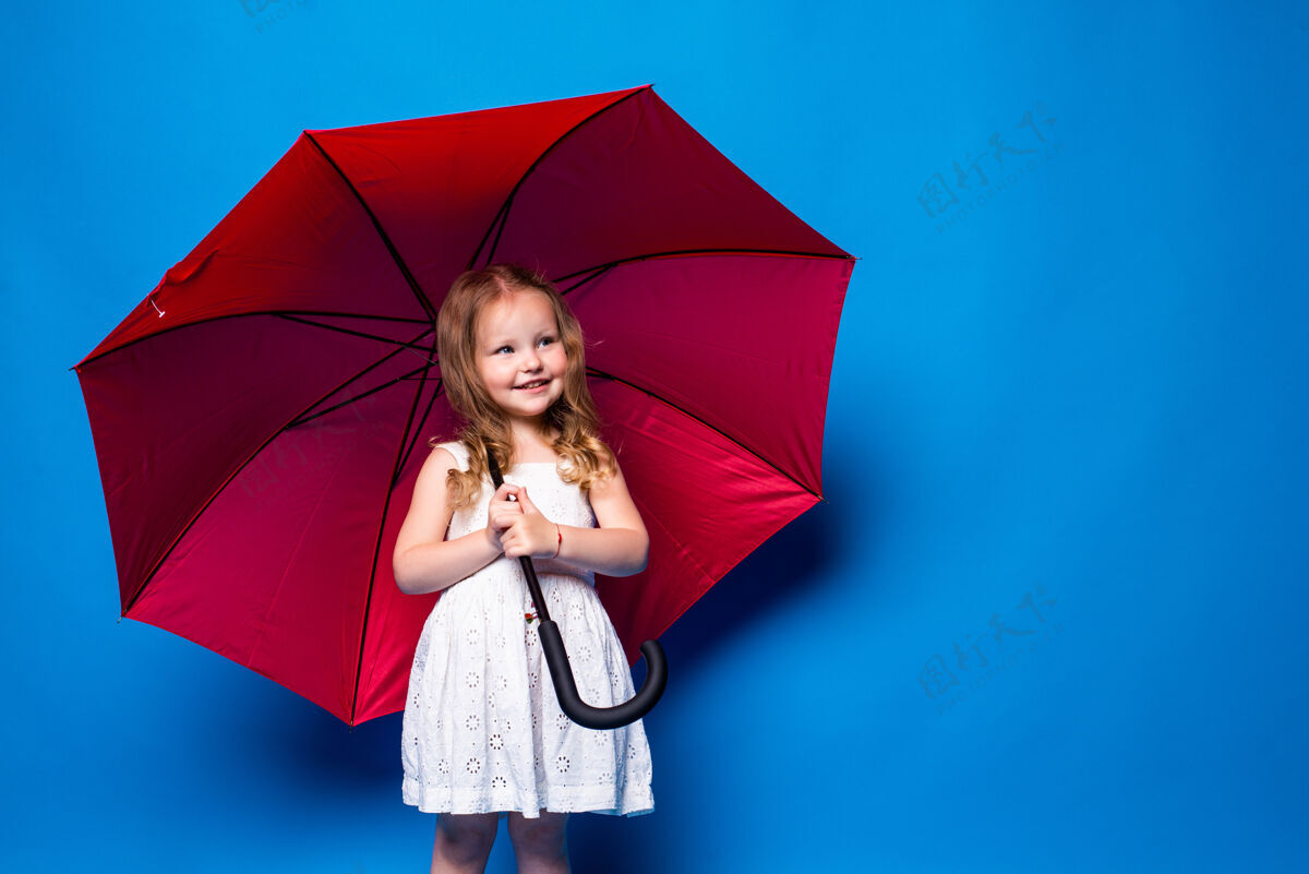 雨快乐的小女孩拿着红伞在蓝色的墙上摆姿势伞雨白裙子