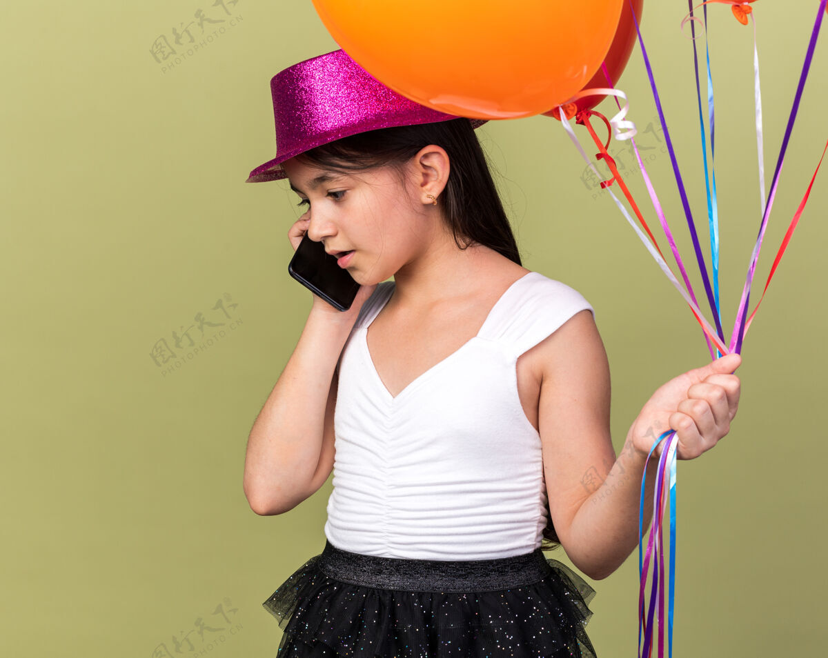 惊喜惊讶的年轻白人女孩 戴着紫色派对帽 手里拿着氦气球 在橄榄绿的墙上讲着电话 墙上留着复印空间帽子白种人气球