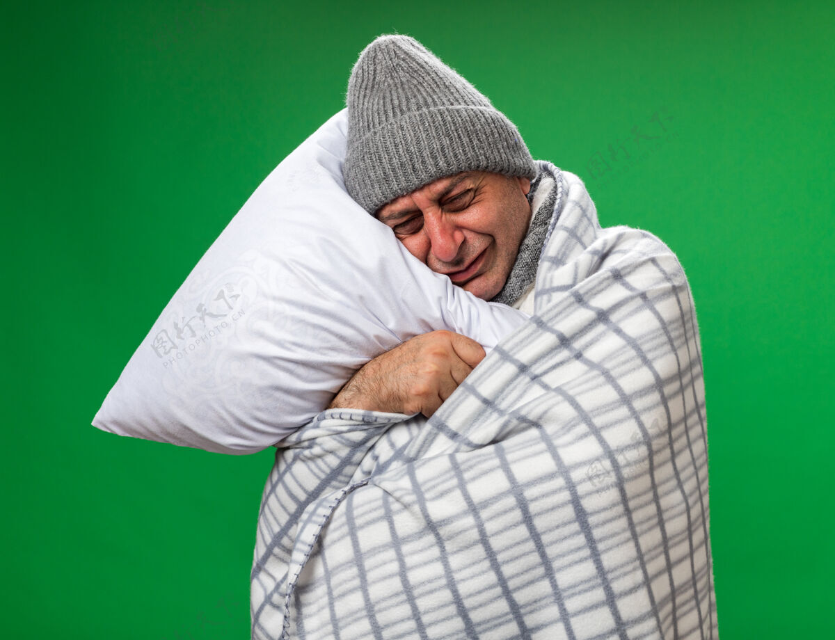 哭哭泣的成年病高加索男子脖子上围着围巾 戴着格子布包裹的冬季帽子 抱着头枕在枕头上 隔离在绿色墙壁上 留着复制空间大人围巾围着