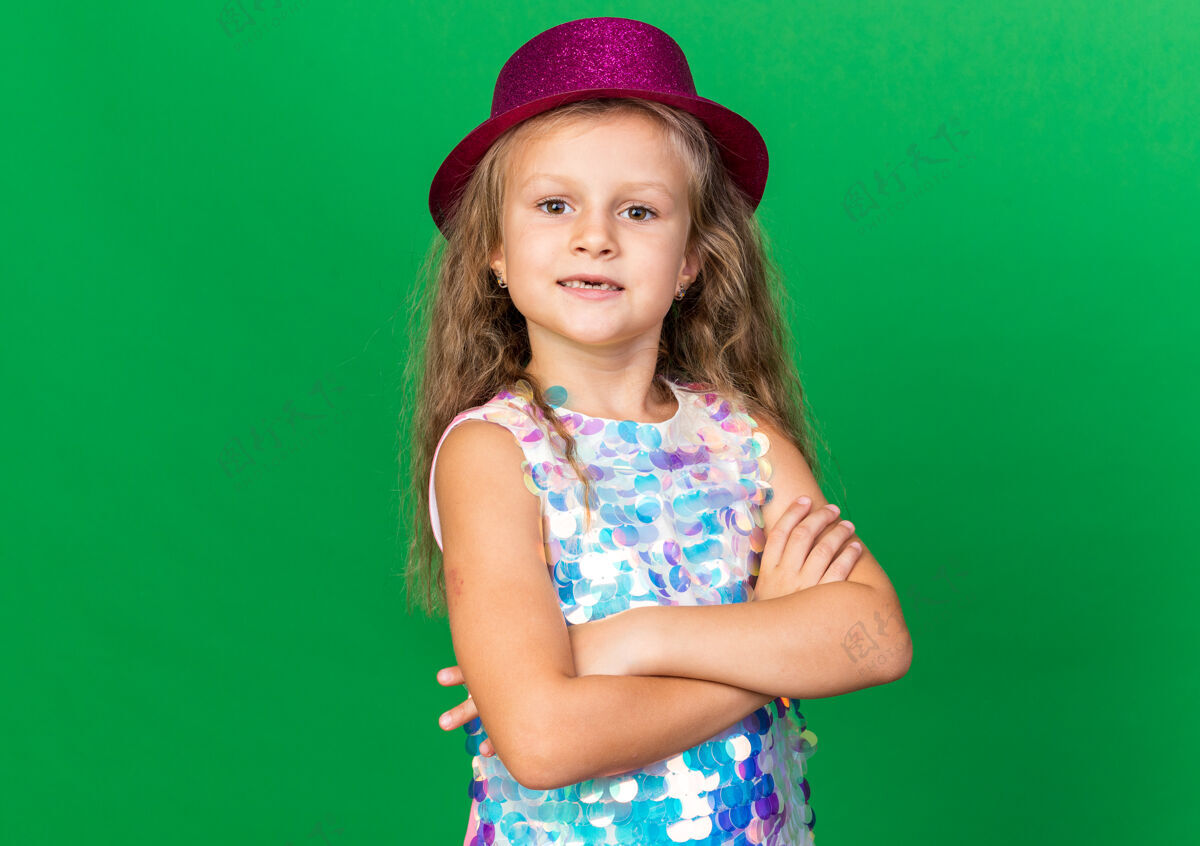 站自信的金发小女孩 戴着紫色派对帽 双臂交叉 站在绿色的墙上 留着复制空间胳膊金发帽子
