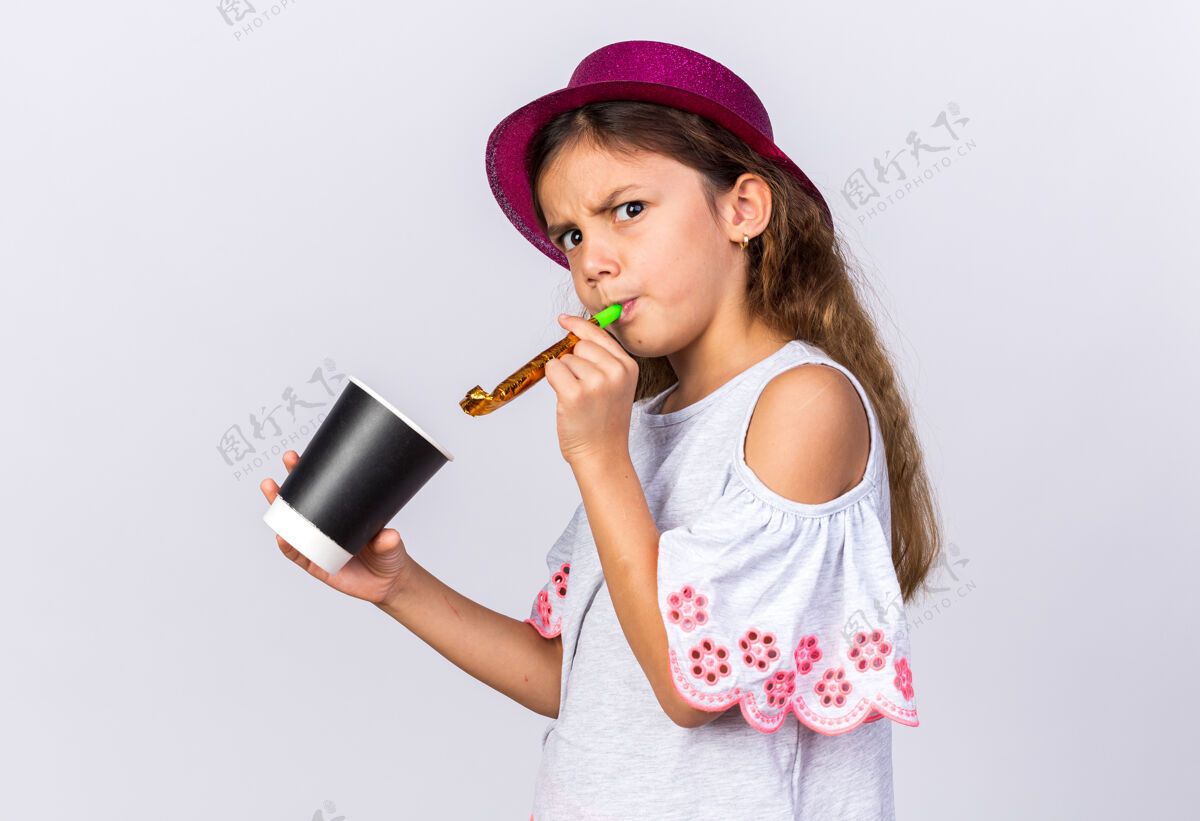 哨子恼怒的小白种女孩戴着紫色的派对帽吹着派对哨子 手里拿着纸杯孤立地放在白色的墙上 留着复印空间恼火生日举着