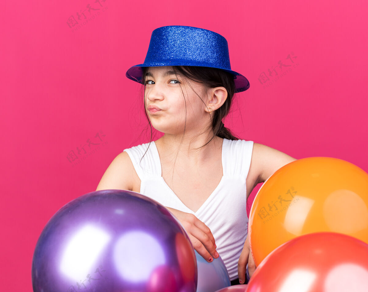 孤立恼怒的年轻高加索女孩戴着蓝色的聚会帽站在粉红色的墙上 氦气球被隔离在复制空间帽子站立气球