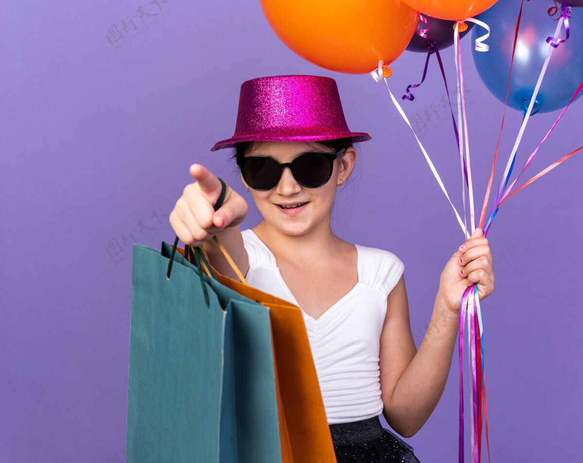 眼镜戴着太阳眼镜 戴着紫色派对帽 拿着氦气球和购物袋的年轻白种人女孩高兴地站在紫色墙壁上 带着复制空间 向前看包指向帽子