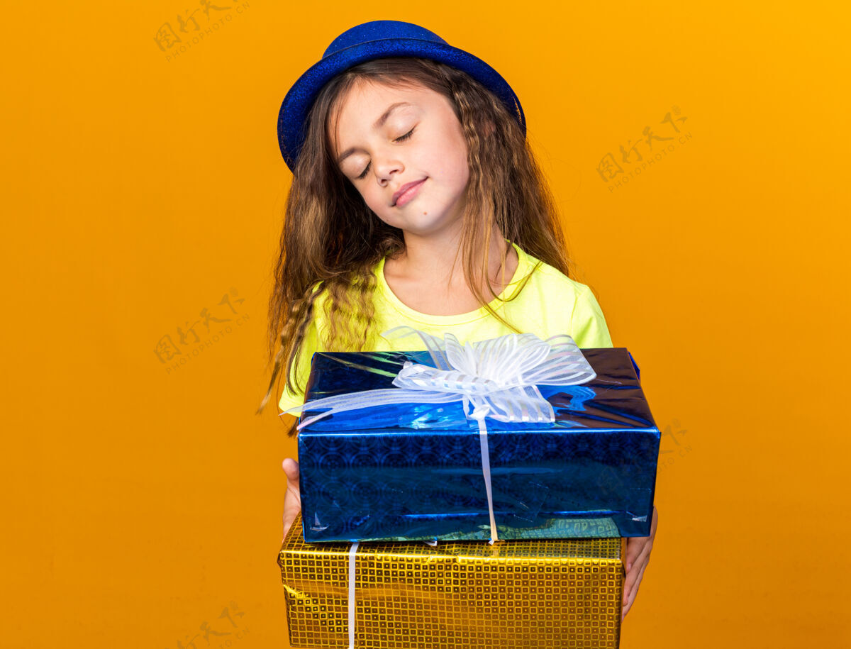 帽子可爱的白人小女孩戴着蓝色派对帽 闭着眼睛站着 手里拿着礼品盒 隔离在橙色的墙上 还有复印空间抱着站着孤立的