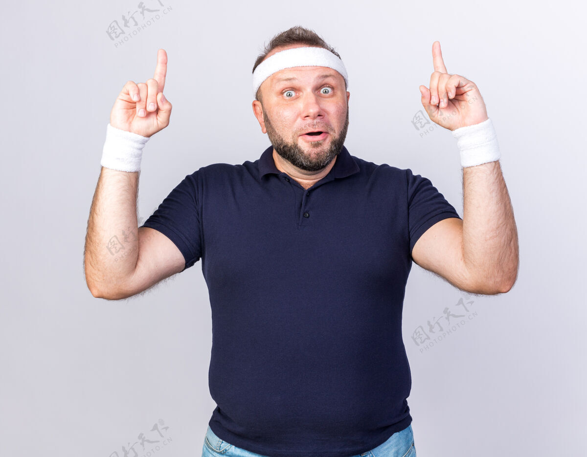指向惊讶的成年斯拉夫运动男子戴着头带和腕带指向隔离的白色墙壁与复制空间男人成人惊喜