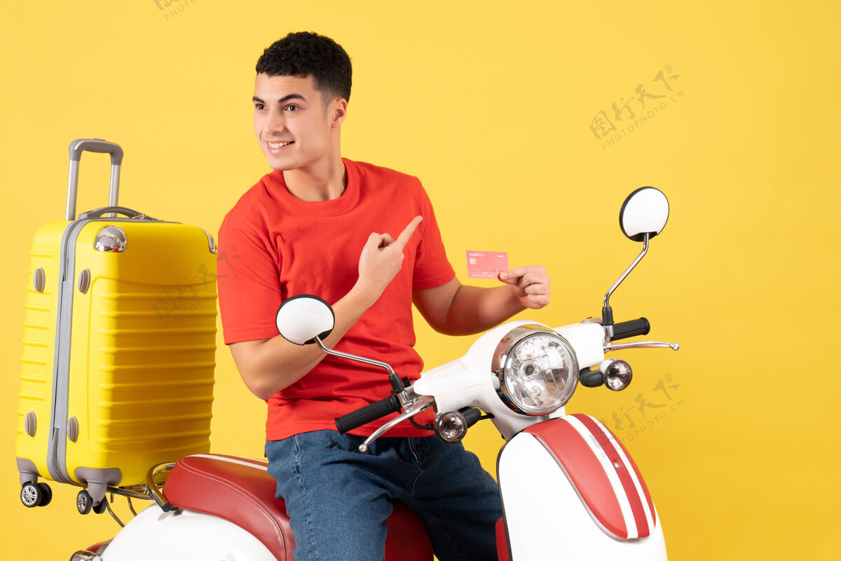男人正面图微笑的年轻人穿着红色t恤 骑着轻便摩托车 手里拿着信用卡人前面持有