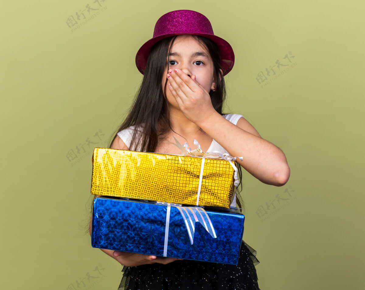 帽子焦虑的年轻白人女孩 戴着紫色派对帽 手放在嘴上 手拿着礼盒 孤立地放在橄榄绿的墙上 留着复印空间女孩橄榄孤立