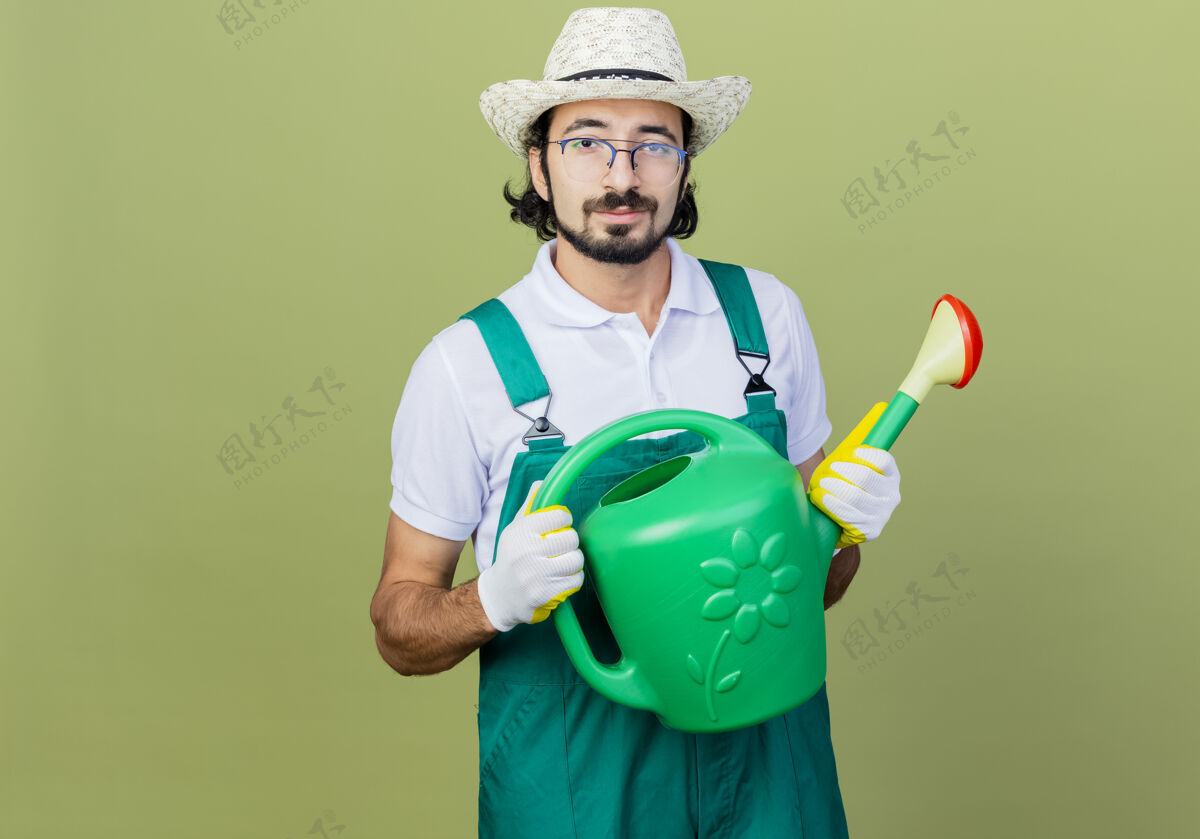 微笑年轻的留着胡须的园丁穿着连体衣戴着帽子拿着浇水罐看着前面微笑着自信地站在淡绿色的墙上人浇水市民