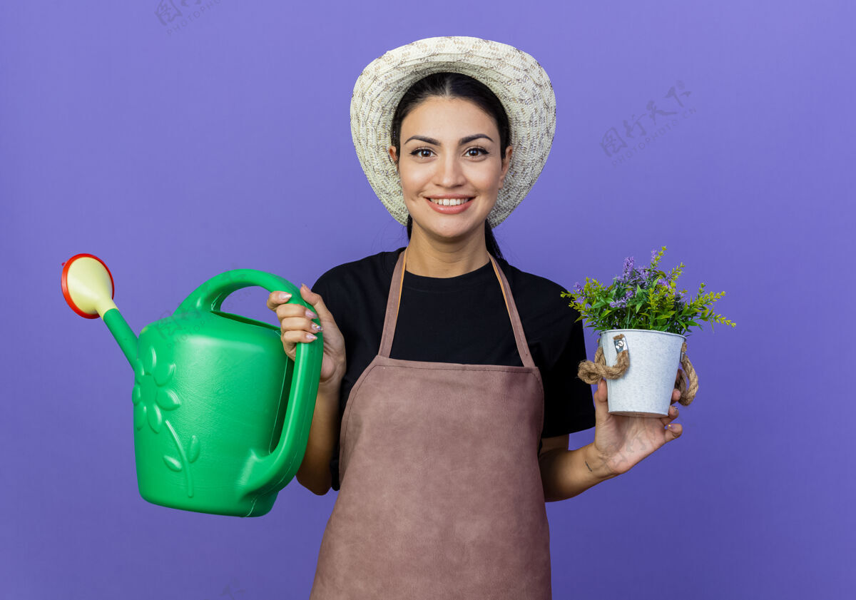 女人年轻漂亮的女园丁围着围裙 戴着帽子 手里拿着水罐和盆栽植物 站在蓝色的墙上 面带微笑地看着前面脸衣服壶