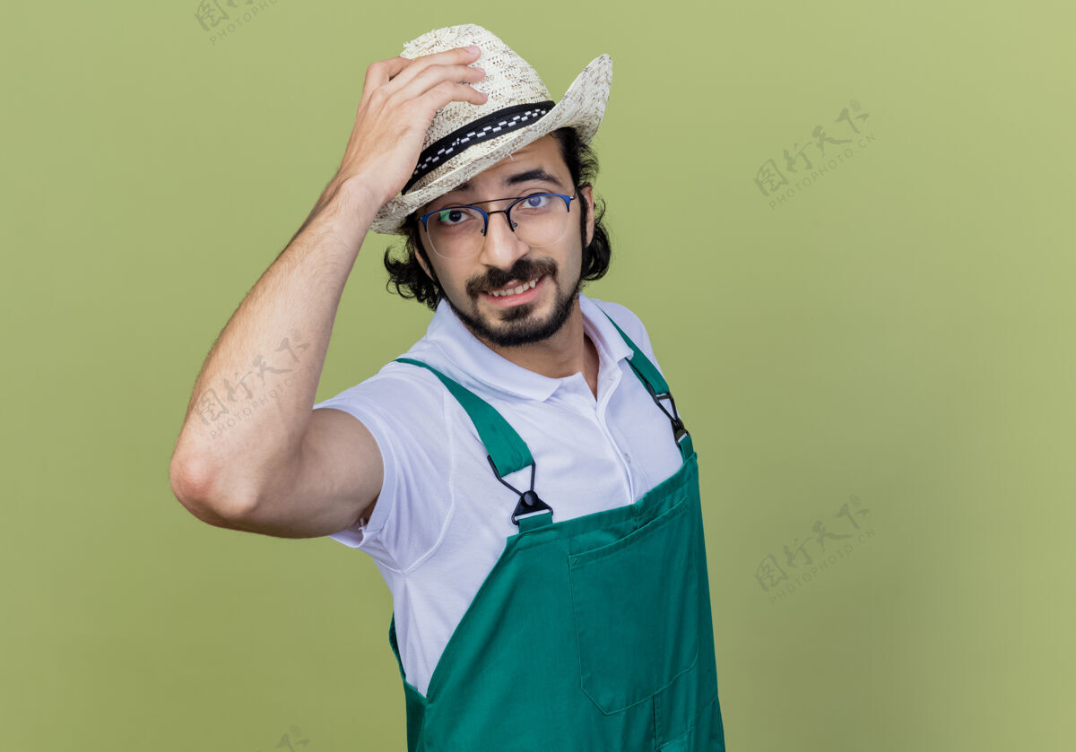 光年轻的留着胡须的园丁 穿着连体衣 戴着帽子 微笑着看着前面 站在浅绿色的墙上 抚摸着他的帽子脸人市民