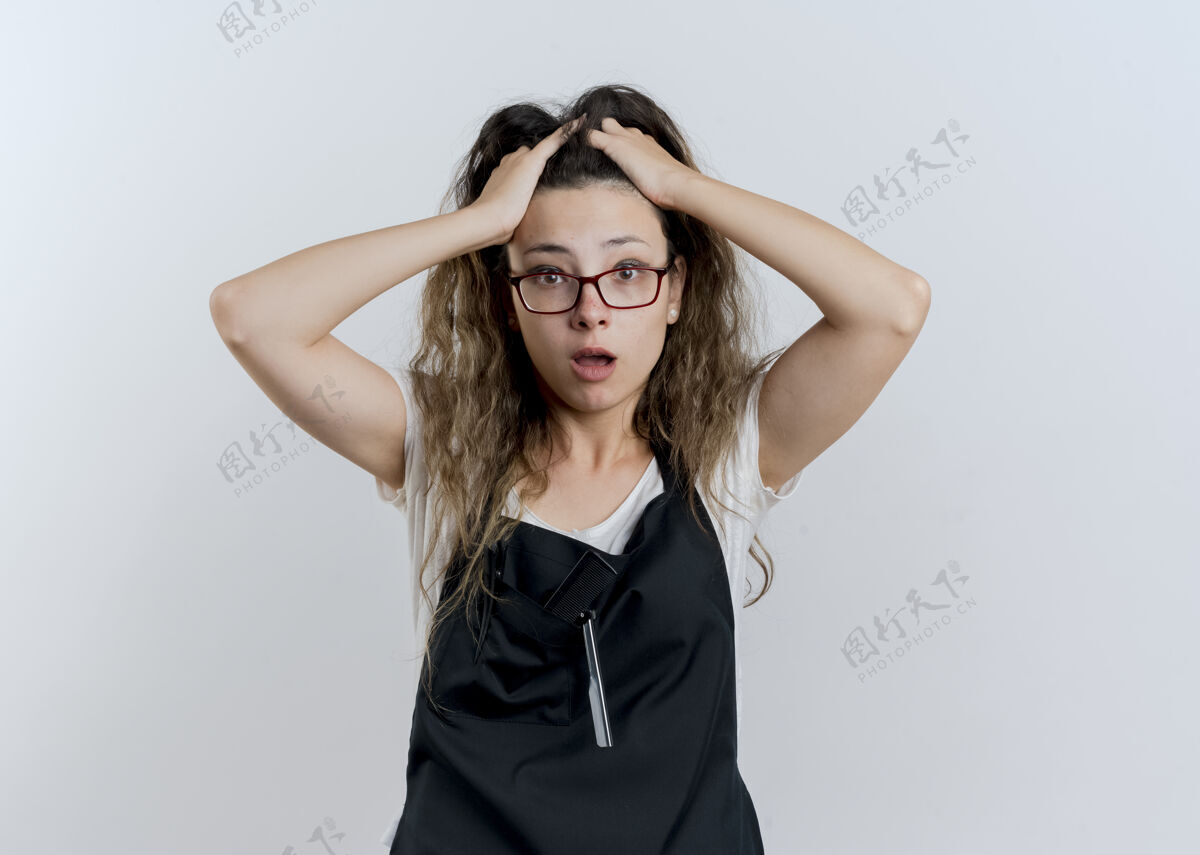 衣服穿着围裙的年轻专业美发师看起来很沮丧 站在白色的墙上拉着她的头发表情专业姿势