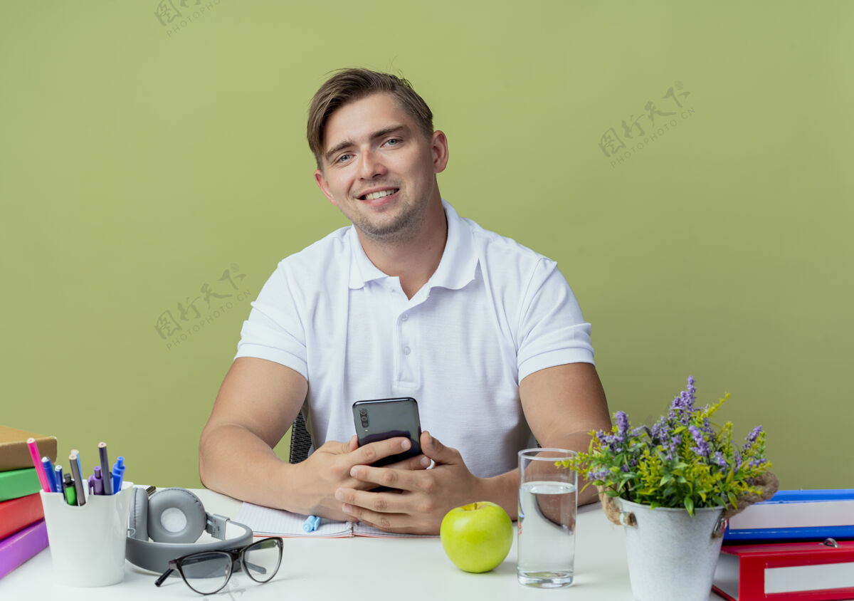 坐着微笑的年轻英俊的男生坐在书桌旁 手里拿着学习用具 手里拿着橄榄绿的电话抱着学校绿色