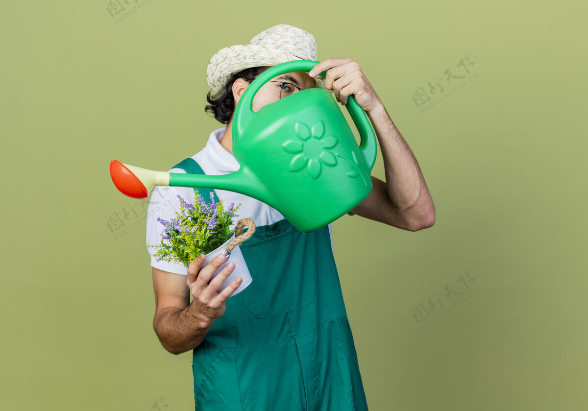 感觉年轻的留着胡子的园丁 穿着连体衣 戴着帽子 手里拿着浇水罐 脸上藏着盆栽植物 站在淡绿色的墙上站着连身衣市民