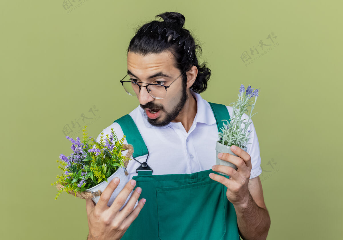 光年轻的留着胡须的园丁 穿着连体衣 手里拿着盆栽植物 站在浅绿色的墙上 困惑地看着它们 试图做出选择花园选择年轻