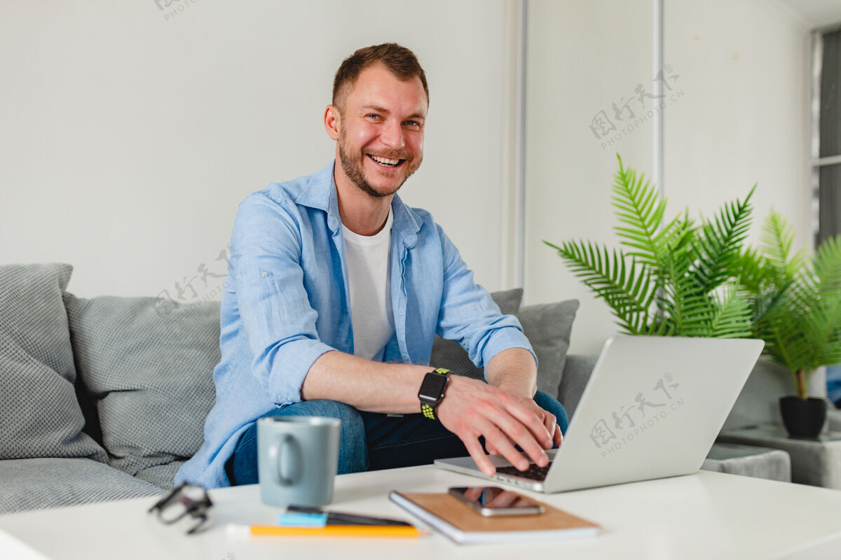 人穿着衬衫的微笑男人在家里的沙发上放松地坐在桌子旁 在家里用笔记本电脑上网笔记本电脑男性室内
