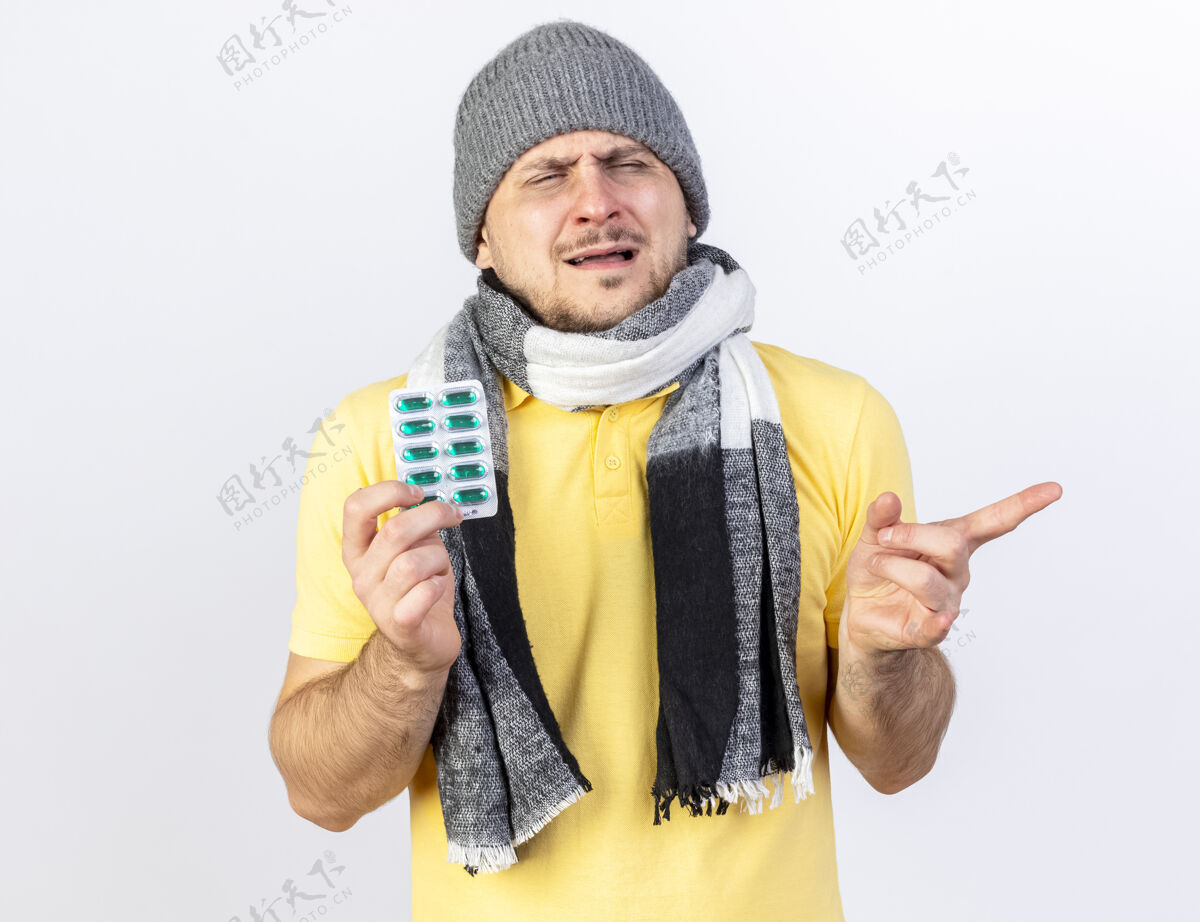 冬天年轻的金发病患戴着冬天的帽子和围巾 拿着一包药片 指着隔离在白墙上的一边持有点穿着