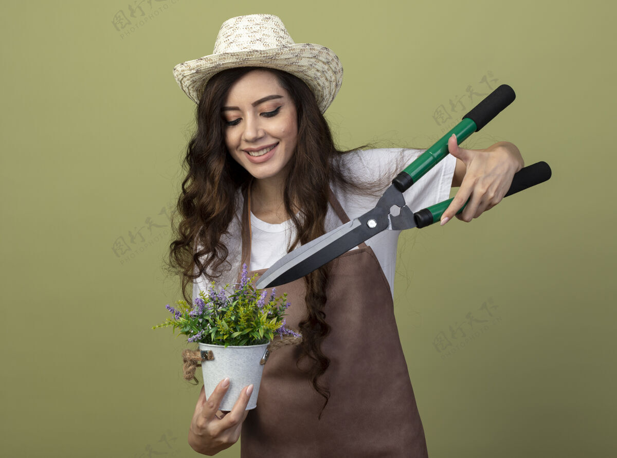 表情身着制服 戴着园艺帽 面带微笑的年轻女园丁把园艺剪举在橄榄绿墙上隔离的花盆里的花朵上剪子年轻花