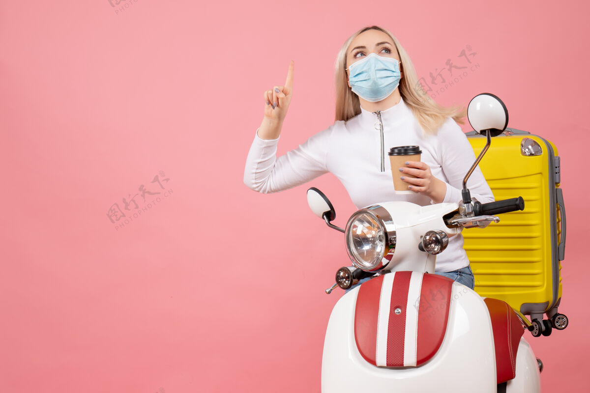 前面正面图：戴着面罩的年轻女士 骑着轻便摩托车 黄色手提箱指着天花板 手里拿着咖啡杯小姐手提箱运动员