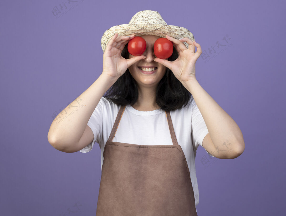眼睛微笑着的年轻黑发女园丁穿着制服 戴着园艺帽 在紫色的墙上用西红柿遮住眼睛帽子黑发花园