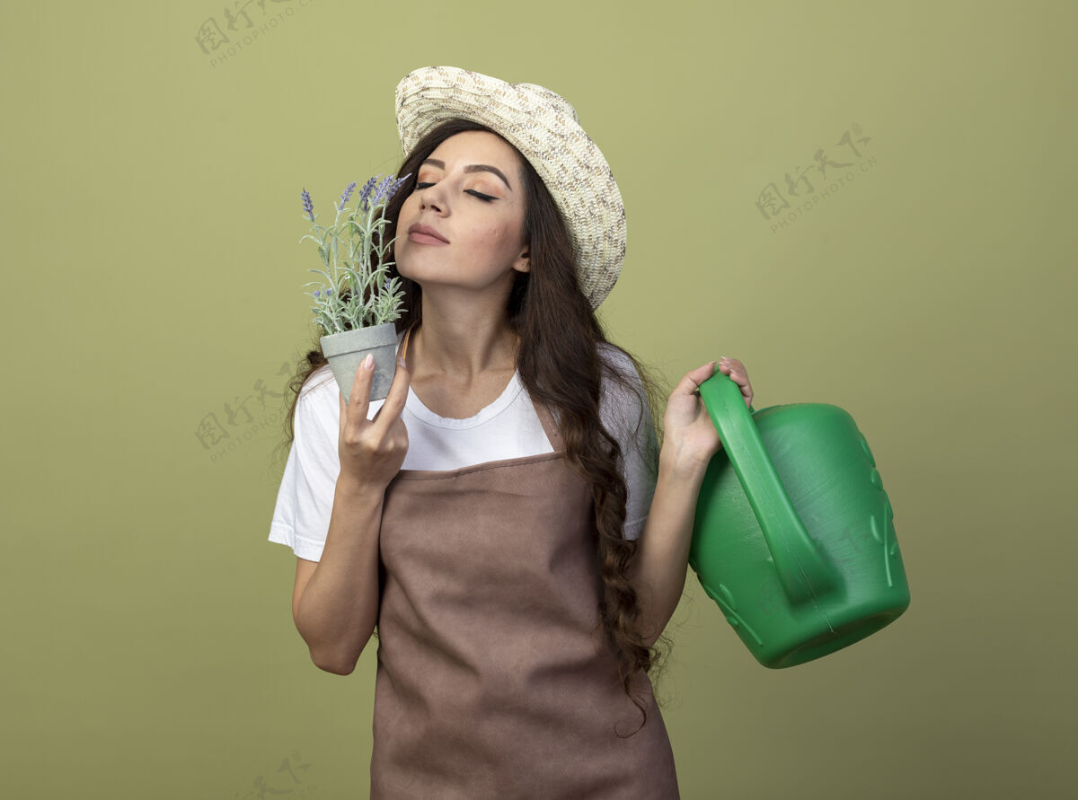 浇水穿着制服 戴着园艺帽的年轻女园丁高兴地拿着喷壶 嗅着橄榄绿墙上孤立的花盆里的花制服年轻帽子