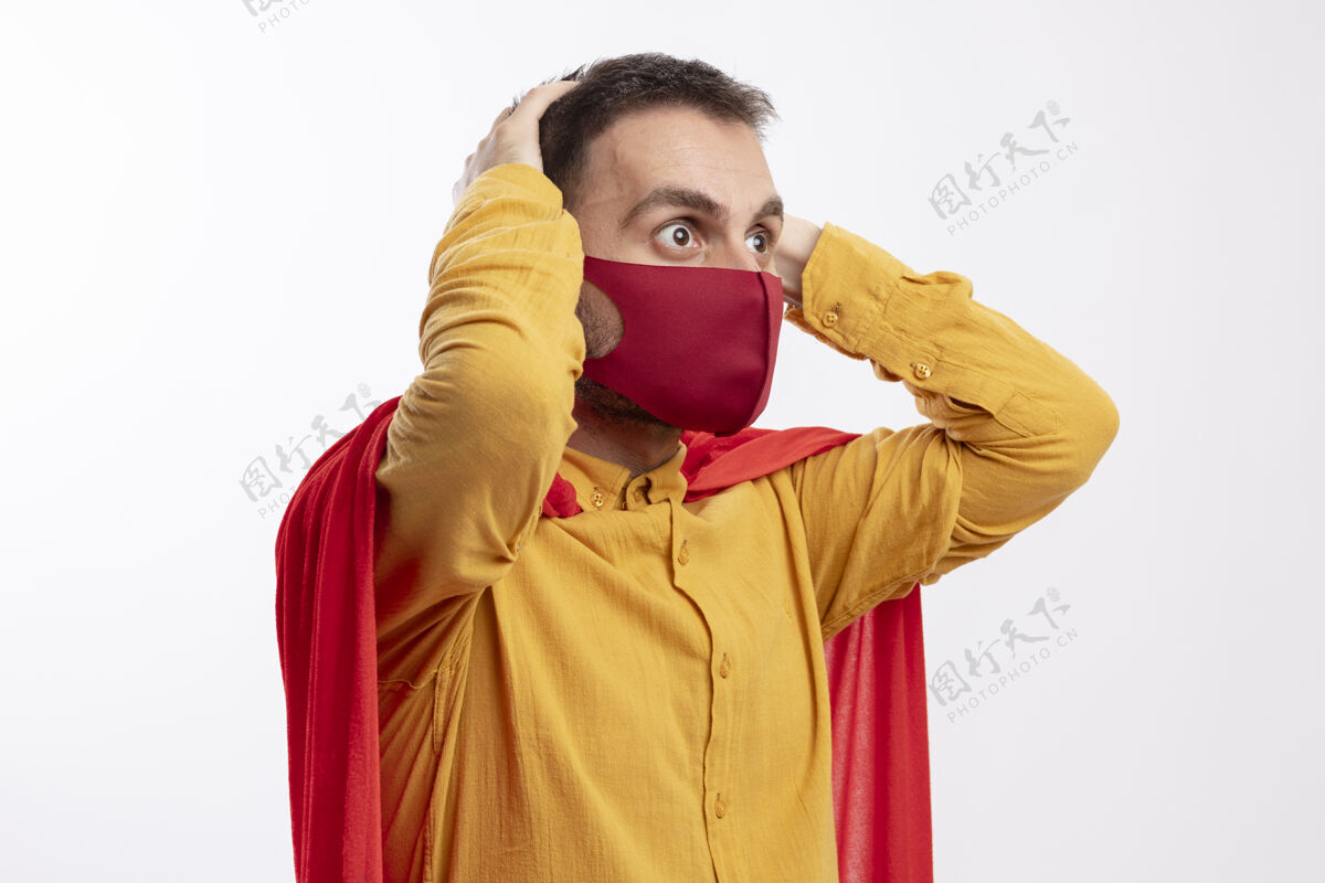 面具焦虑的超级英雄身穿红色斗篷 戴着红色面具 双手放在头上 看着隔离在白墙上的一面公民姿势焦虑