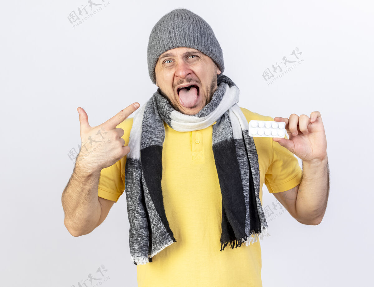 壁板恼怒的年轻金发病男子戴着冬天的帽子和围巾 手里拿着一包药片 指着隔离在白墙上的一边脸人表情