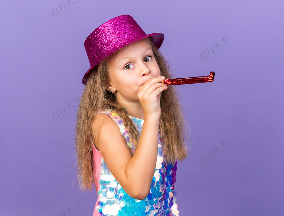 帽子可爱的金发小女孩戴着紫罗兰色的派对帽吹着派对哨子 在紫色的墙壁上看着与世隔绝的复制空间金发孤立哨子