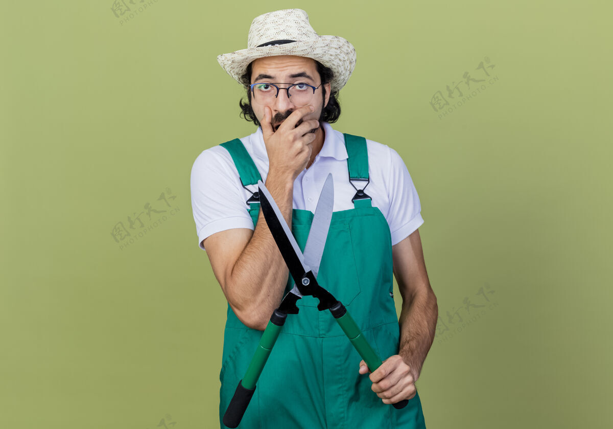 人年轻的留着胡须的园丁 穿着连体衣 戴着帽子 手持树篱剪 站在浅绿色的墙上 看上去既惊讶又惊奇市民篱笆惊人