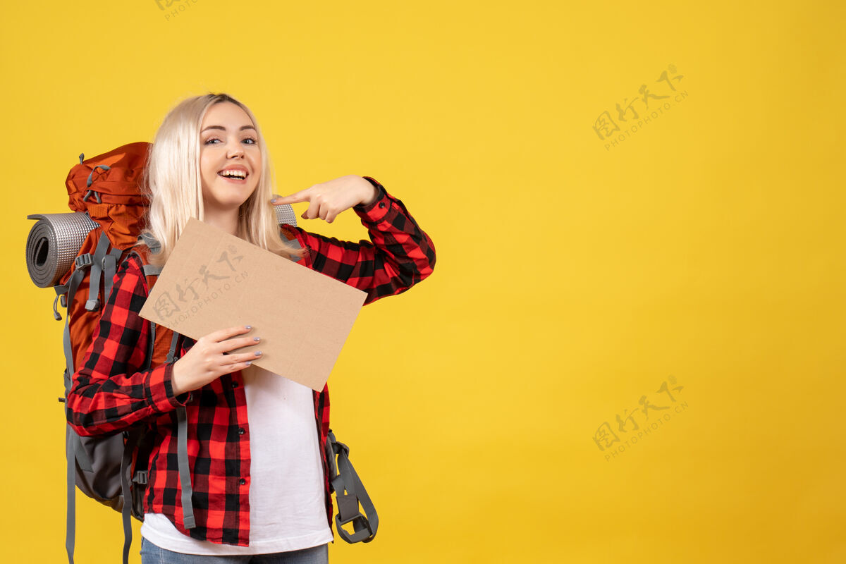 快乐正面图微笑的金发女旅行者背着她的背包拿着纸板站在黄色的墙上成人背包女士