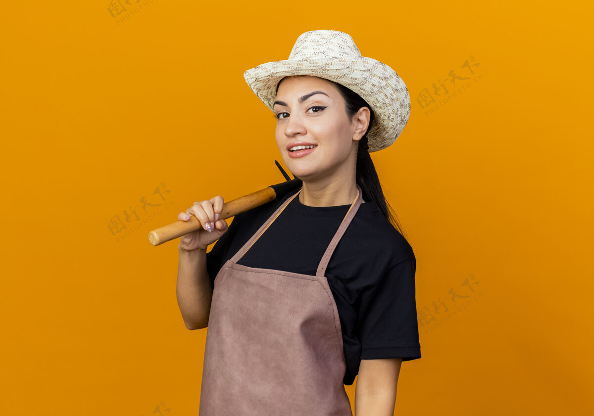 站着年轻漂亮的女园丁围着围裙 戴着帽子 拿着小耙子 站在橙色的墙上 面带微笑地看着前面抱着表情女人