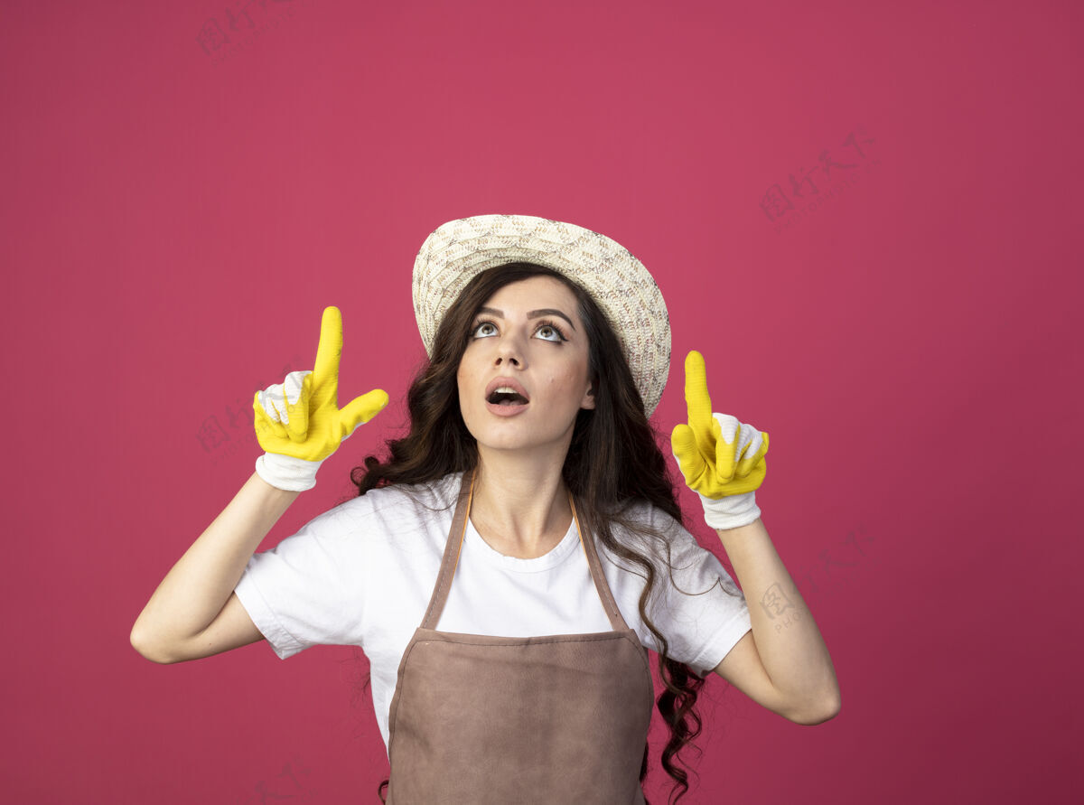 女人穿着制服 戴着园艺帽和手套的年轻女园丁惊讶地看着粉色的墙上孤零零地指了指园艺人脸