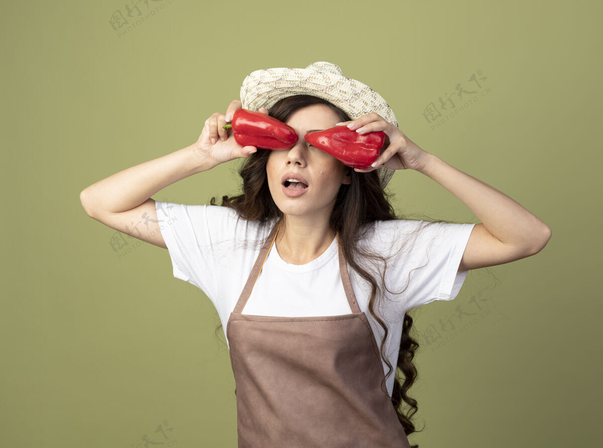 橄榄穿着制服 戴着园艺帽的年轻女园丁惊讶地用橄榄绿墙上隔离的红辣椒遮住眼睛封面胡椒眼睛