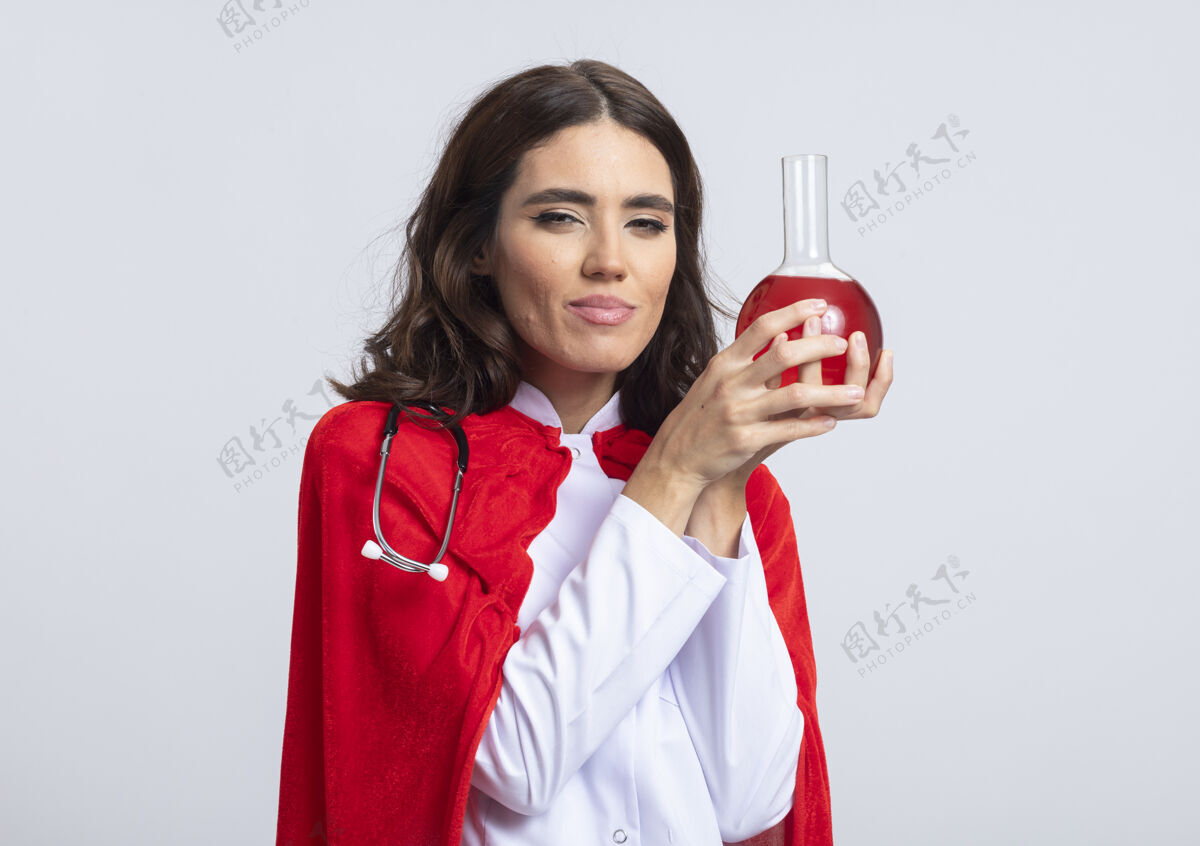 姿势身着医生制服 身穿红色斗篷 手持听诊器的快乐女超人将红色化学液体装在隔离在白墙上的玻璃瓶中请玻璃女孩