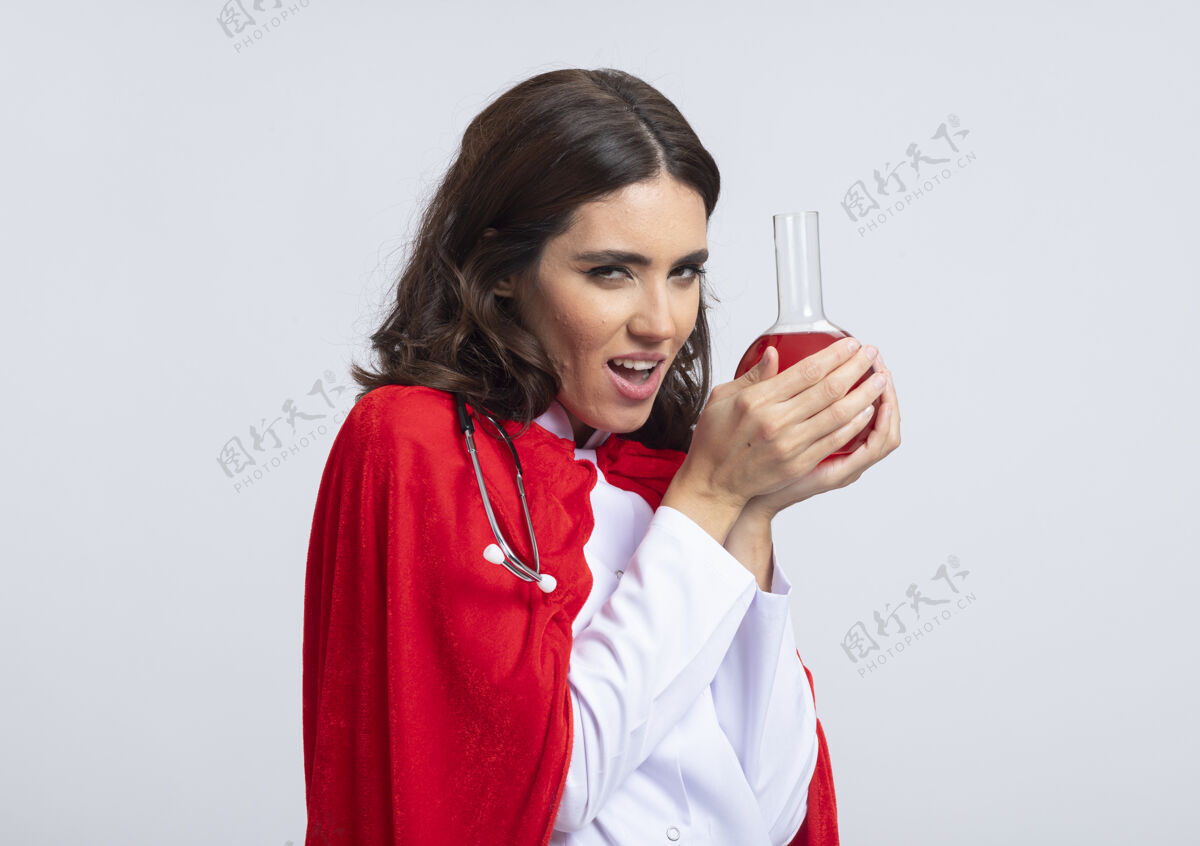 化学品邪恶的快乐女超人穿着医生制服 戴着红色斗篷和听诊器 把红色的化学液体装在隔离在白墙上的玻璃瓶中制服人玻璃